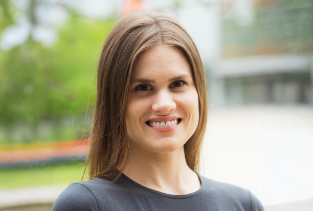 Smiling white woman with light brown hair wearing a gray top; outdoor background is blurred.
