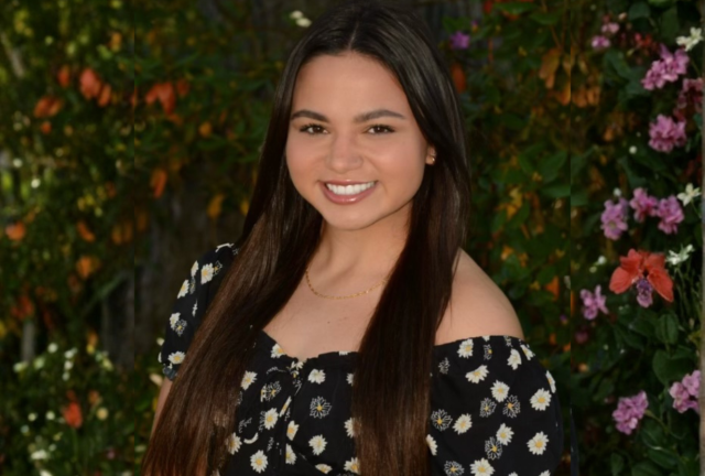 Smiling white female with long brown hair wearing a black top with yellow and white fllowers; blurred background of flowering trees.