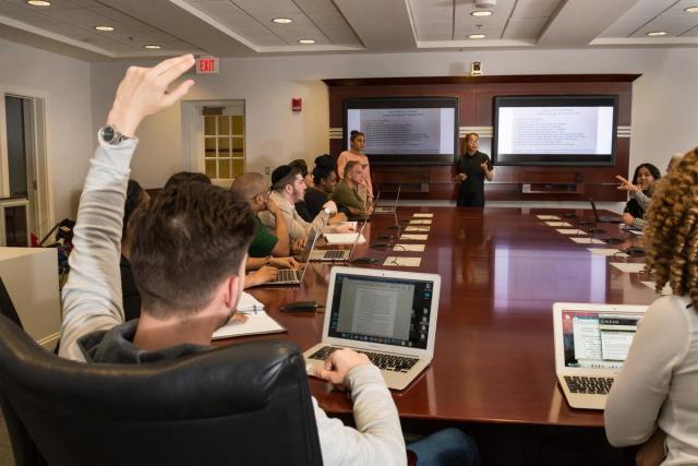 Students sitting around a large wooden conferenceroom table with laptops; one has their arm up.