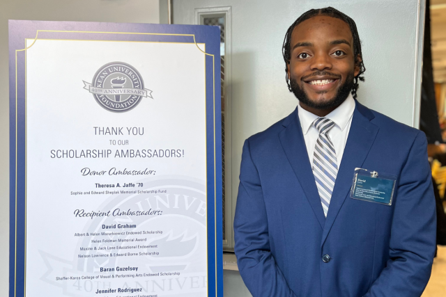 Smiling young Black male student in a blue suit and grey striped tie stands next to a sign announcing his scholarships.