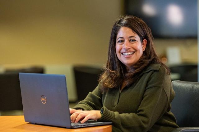 Smiling female with brown hair wearing a green sweater looks up from her laptop.
