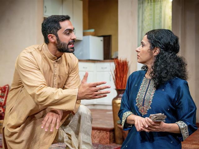 Two people talk in a living-room stage set. One person, wearing traditional clothing, gestures with an open hand, while the other—also in traditional attire—holds a stack of papers or money. A kitchen area is visible in the background. 