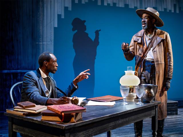 Two performers interact in a stage setting. One sits at a desk covered with books and papers, while the other stands wearing a long coat and hat. A shadow of the standing figure is cast prominently on the wall behind them. 