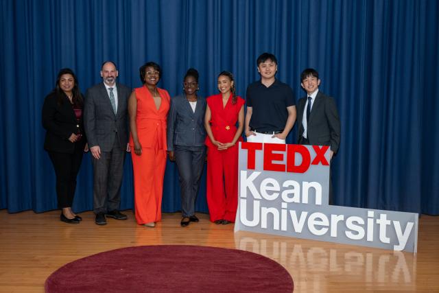 men and women lined up on a stage next to a sign reading TedxKean University