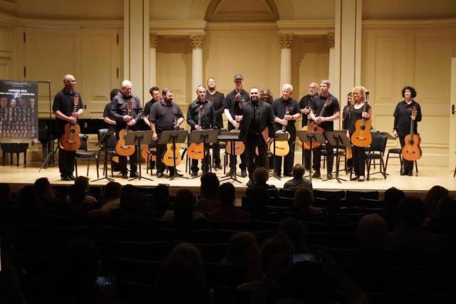 A group of musicians on stage, members of the New Jersey Guitar Orchestra, one man in a suit is standing up and the shot is from audience view