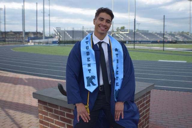 Man standing outside wearing graduation gown in front of athletic fields
