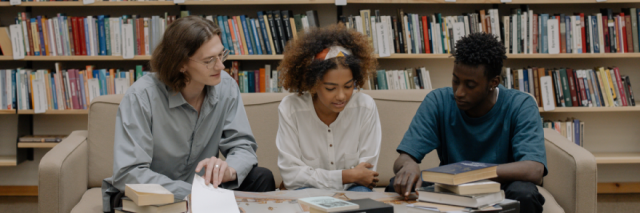3 students are seated on a sofa next to each other, looking at the study materials on the table in front of them.