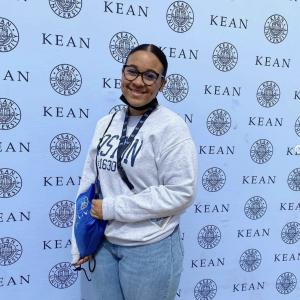 A young woman in a gray sweater with glasses smiling in front of a backdrop with the Kean logo on it.